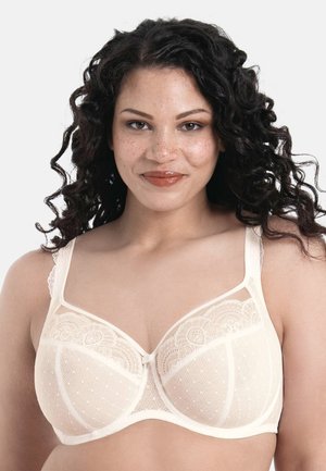 Vrouw met krullend donker haar lacht, draagt een gebroken-witte kanten beha met verstelbare bandjes tegen een effen lichte achtergrond.