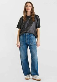 Donna con lunghi capelli castani che indossa una maglietta nera a maniche corte larga, jeans blu larghi con grandi tasche anteriori e sneakers bianche, in piedi su uno sfondo bianco.