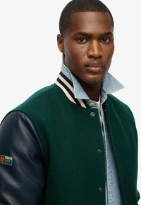 Groene wolmix varsityjack met marineblauwe leren mouwen, gestreepte kraag en drukknoopsluiting, gedragen over een lichtblauw denimshirt.