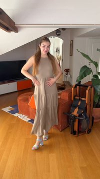 Νεαρή γυναίκα με μακριά μαλλιά φορεί μπεζ αμάνικο φόρεμα και ανοιχτό μπλε τακούνια, στέκεται σε ένα σύγχρονο σαλόνι με ξύλινο πάτωμα και φυτά.