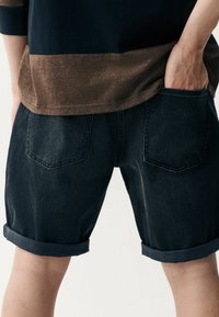Dunkelblaue Denim-Shorts mit umgeschlagenen Säumen, ausgestattet mit zwei Gesäßtaschen und einer glatten Textur. Das Oberteil hat ein braunes, horizontales Streifenmuster.