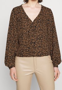 Fließende schwarze Bluse mit kleinem orangefarbenem Blumenmuster, V-Ausschnitt und langen Ärmeln. Kombiniert mit beigen, taillierten Hosen. Dezente Knöpfe an der Vorderseite.