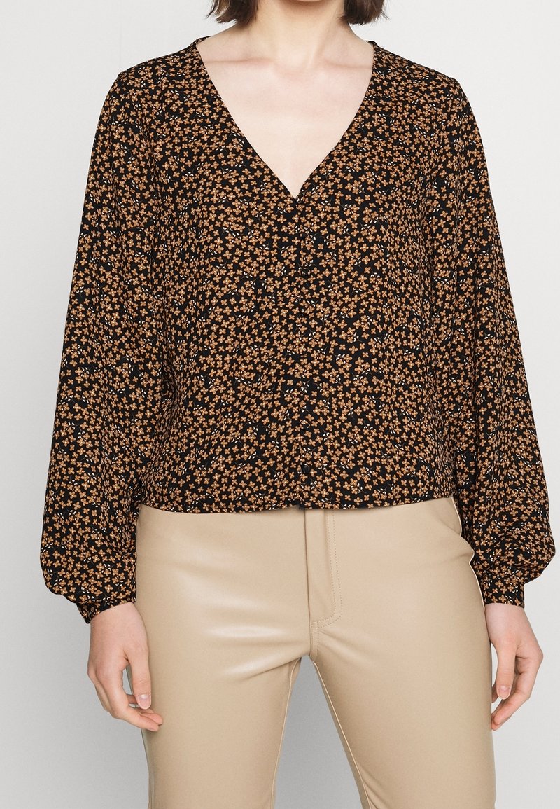 Fließende schwarze Bluse mit kleinem orangefarbenem Blumenmuster, V-Ausschnitt und langen Ärmeln. Kombiniert mit beigen, taillierten Hosen. Dezente Knöpfe an der Vorderseite.