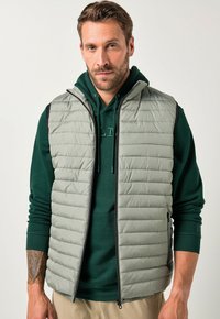 Gepolsterte Weste in Hellgrau mit hohem Kragen, mit einem Reißverschluss vorne und schwarzen Akzenten auf den Schultern. Getragen über einem grünen Hoodie.
