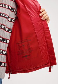 Czerwona, ocieplana kurtka z gładkim nylonowym wnętrzem, z metką informującą, że zawiera 100% przetworzonego poliestru. Widoczny sznurek ściągający i zamek błyskawiczny.