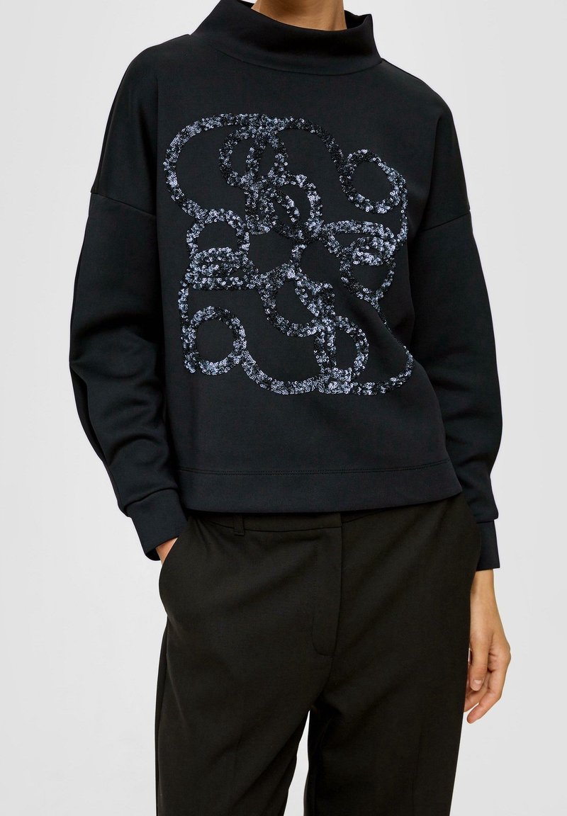 Sudadera negra con cuello tipo alto, que presenta un diseño de lazos de lentejuelas en tonos de azul. Ajuste casual con puños y dobladillo acanalados.