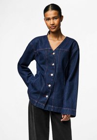 Traper jakna tamnoplave boje s V-izrezom, kopčanjem na gumbe i crvenim šavovima. Ima duge rukave i bočne džepove.