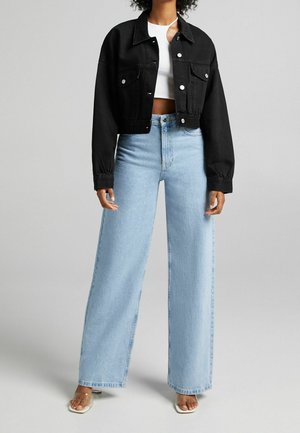 Person iført en sort cropped denimjakke over en hvid crop top, lyseblå vide jeans og hælede sandaler med klare remme.