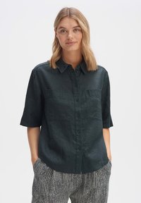 Camicia a maniche corte verde scuro con bottoni e due tasche sul petto, realizzata in un tessuto testurizzato. Abbinata a pantaloni neri e bianchi con motivo.
