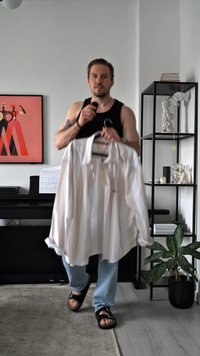 White striped shirt held by a person, featuring a collar and long sleeves. The fabric appears lightweight. Background includes a black shelf and a plant.