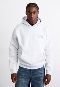 Gray hooded sweatshirt with a front pocket and embroidered logo on the left chest. Worn with blue denim jeans featuring a straight cut.