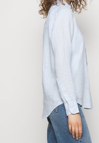 Kvinna med lockigt hår som bär en ljusblå långärmad skjorta och blå jeans, stående sidledes mot en enkel bakgrund.