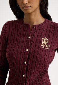 Vīna sarkana džempera jaka ar pīnītas adīšanas rakstu, zelta krāsas pogām un kreisajā krūtīs izšūtu krēmkrāsas logotipu.