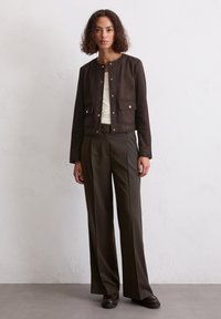 Braune Wildlederjacke mit silbernen Knöpfen und zwei Fronttaschen. Getragen über einem weißen Hemd mit maßgeschneiderten olivgrünen weiten Hosen und braunen Schuhen.