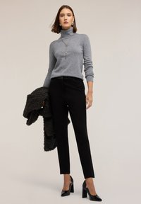 Grijze coltrui, strak zittende zwarte broek en zwarte hoge hakken. Model houdt een gestructureerde zwarte jas over haar arm.