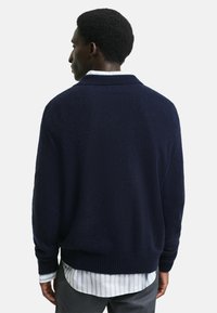 Granatowy sweter dzianinowy o luźnym kroju, z prążkowanymi mankietami i dołem oraz kołnierzem ze stójką. Noszony na koszuli w paski z kołnierzykiem. Miękka tekstura.