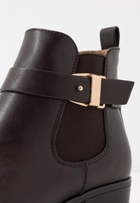 Botín marrón hasta el tobillo hecho de cuero sintético con una textura suave, con una correa con hebilla en tono dorado y paneles laterales elásticos para facilitar su uso.