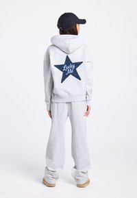 Grijze hoodie met een marineblauwe ster en de tekst "Lucky Me" op de achterkant, gecombineerd met bijpassende grijze joggingbroek en beige schoenen.