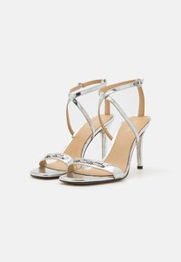 Sandale cu toc înalt din metalic argintiu, cu un design cu barete încrucișate, vârf deschis și o finisare texturată. Prezintă un toc stiletto elegant și închidere cu cataramă.