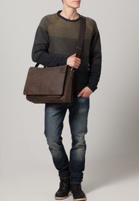 Brun læder messenger taske med en fold-over flap, justerbar skulderrem og subtile stikninger, båret af en person iført en tekstureret sweater.