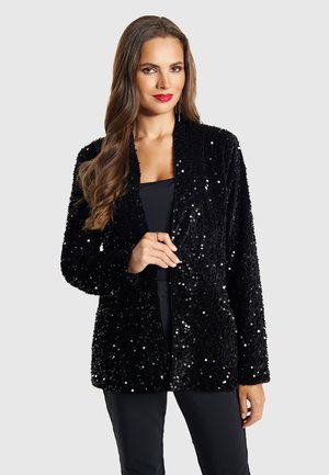 Femeie cu păr lung și castaniu poartă un sacou negru cu paiete peste un top și pantaloni negri, cu ruj roșu și cercei lungi.
