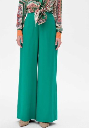 Pantalons larges verts en tissu lisse, taille haute, avec une silhouette fluide. Assortis à un chemisier à motifs.