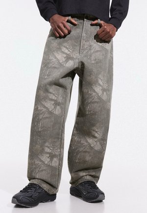 Personne portant un pantalon ample gris tie-dye et des baskets noires, debout les mains dans les poches devant un fond uni.
