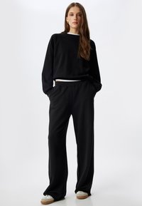 Zwarte sweatshirt met lange mouwen en een losse pasvorm, gecombineerd met zwarte wijde broek. Beide zijn gemaakt van zachte stof en hebben zijzakken.