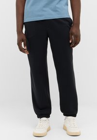 Schwarze, elastische Jogginghosen mit lockerer Passform und gerafften Bündchen. Kombiniert mit weißen Turnschuhen und einem blauen T-Shirt.