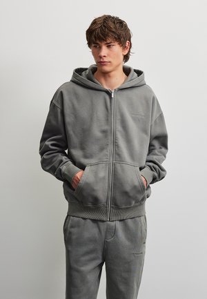 Jeune homme aux cheveux bruns portant un sweat à capuche gris zippé et un pantalon de survêtement assorti, les mains dans les poches du sweat.