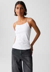 Woman with short dark hair wearing a white camisole and faded black jeans, standing with hands in back pockets against a plain background.