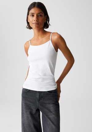 Frau mit kurzem dunklem Haar, die ein weißes Trägertop und farblich verblasste schwarze Jeans trägt, steht mit den Händen in den hinteren Taschen vor einem schlichten Hintergrund.