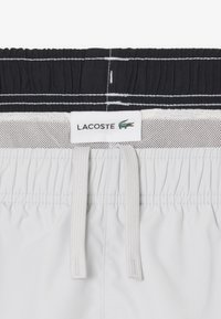 Lacoste zwembroek met een elastische zwart-witte tailleband, lichtgewicht grijs materiaal, vetersluiting aan de voorkant en een zichtbaar logo-label.