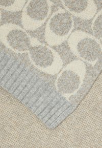 Cachecol cinza de malha com um padrão circular em creme e castanho claro. Textura canelada numa das bordas, material suave e espesso.