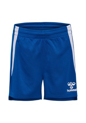 Ung pige står og har blå sportstøj på med hvide og marineblå accenter, hvide sokker og hvide sneakers med sorte chevron-logoer.