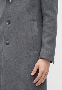 Main droite à côté d’un manteau en laine gris avec trois boutons visibles et une poche verticale fendue sur le devant du manteau.
