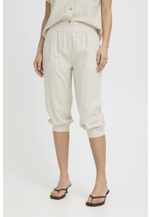 Frau trägt beige gekürzte Hosen mit elastischem Bund und schwarze Sandalen mit Absatz, steht vor einem einfarbigen Hintergrund.