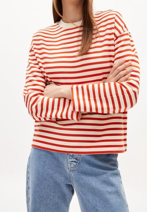 Person iført en langærmet hvid skjorte med orange vandrette striber og lyseblå jeans, med armene krydsede over brystet.