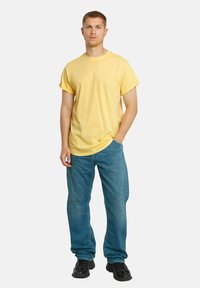 T-shirt di cotone gialla con maniche corte, collo rotondo e vestibilità rilassata, indossata con jeans azzurri chiari e scarpe da ginnastica nere. Design semplice senza motivi.