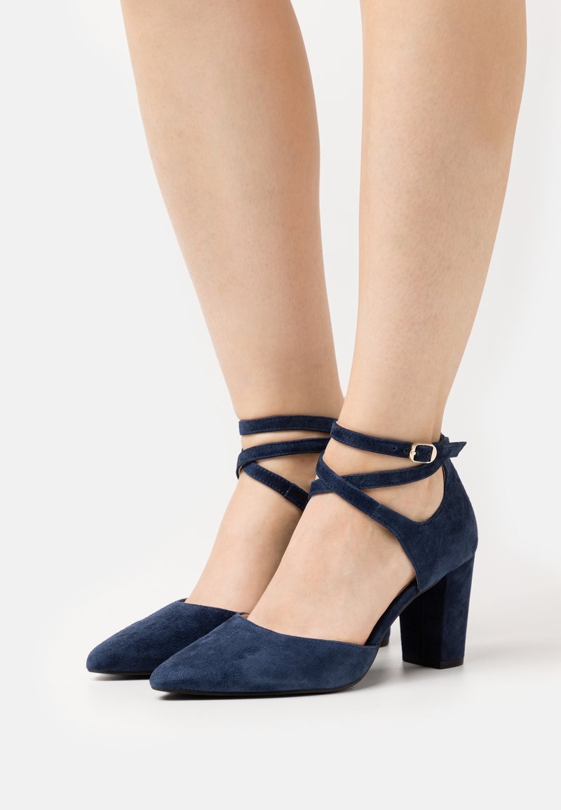 Femme portant des talons blocs en daim bleu marine avec bouts pointus et brides croisées à la cheville avec petites boucles.