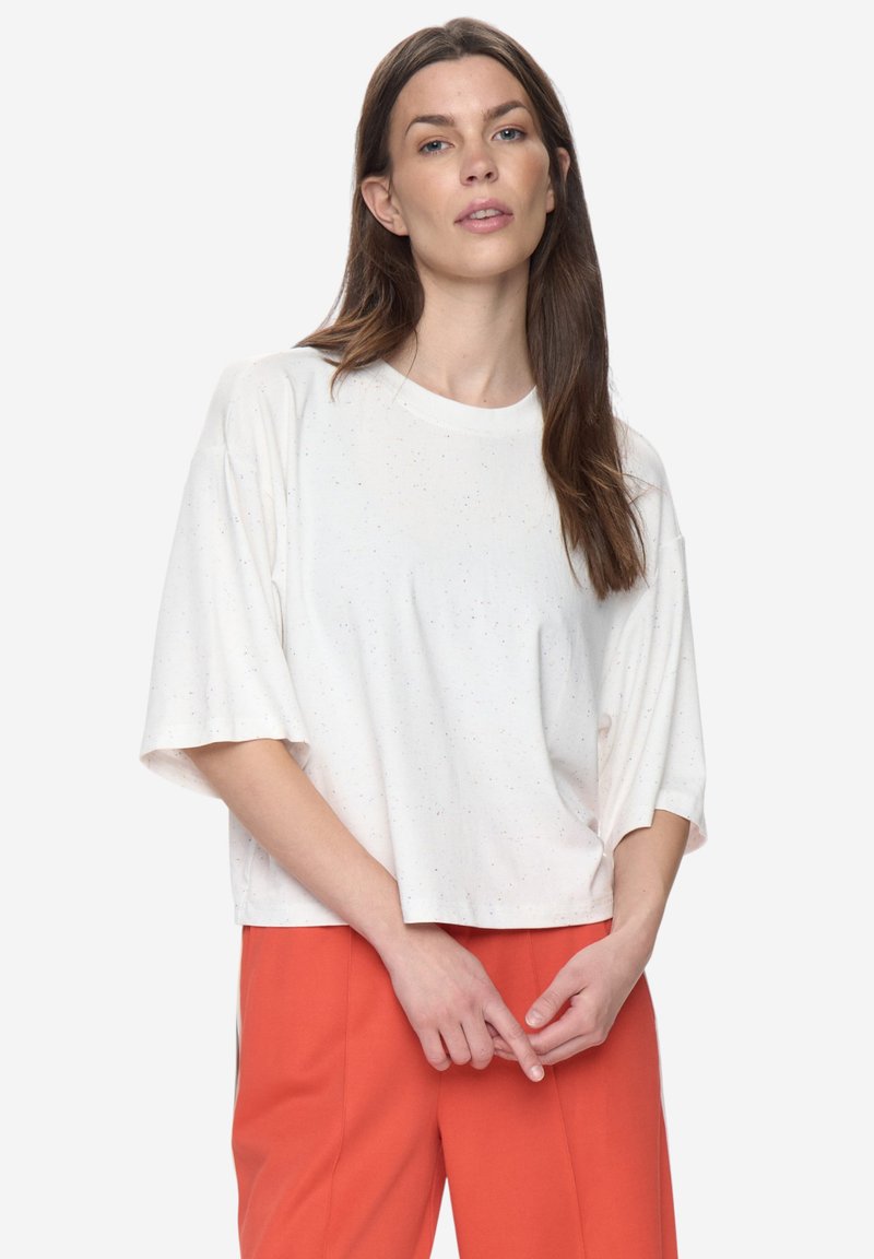 T-shirt blanc à manches courtes avec une coupe décontractée et une texture légèrement mouchetée. Associé à un pantalon orange, mettant en valeur la simplicité du haut.