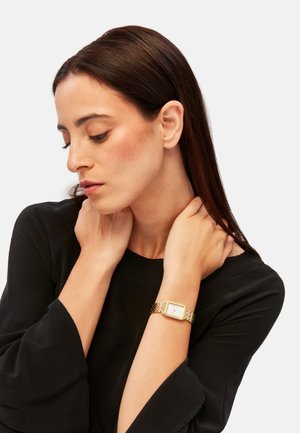Orologio d'oro con quadrante quadrato, bracciale a maglie metalliche e quadrante bianco. Il modello indossa una blusa nera con maniche a campana, le mani appoggiate sul collo.