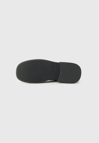 Zapato negro tipo slip-on con suela de goma texturizada, que presenta surcos horizontales para un mejor agarre y una parte superior lisa y redondeada.