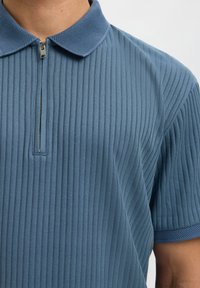Polo bleu côtelé à manches courtes avec un col texturé et une patte de boutonnage partiellement zippée sur la partie supérieure du torse d'une personne.