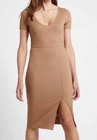 Kobieta w żeńskiej, prążkowanej sukience midi w odcieniu brązowym, z krótkim rękawem i głębokim dekoltem w kształcie litery V, z rozcięciem sięgającym nad kolano, ozdobiona warstwami złotych naszyjników.