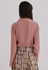 Vrouw met lang, donker haar die een stoffige roze blouse met lange mouwen draagt, ingevoerd in een taille hoge rok met veelkleurige verticale en horizontale strepen.