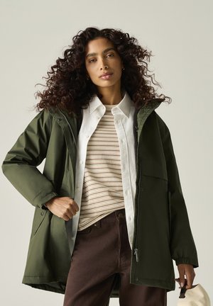Mujer con cabello rizado que lleva una chaqueta con capucha verde, camisa blanca y suéter a rayas, sosteniendo un sombrero beige sobre un fondo liso.