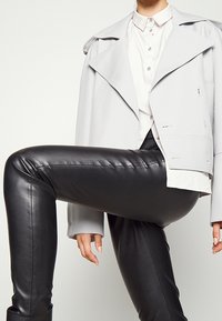 Personne portant une chemise blanche boutonnée, une veste gris clair et un pantalon en cuir noir, avec un genou levé, sur un fond uni.