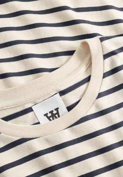 Gros plan sur une chemise en coton crème avec des rayures horizontales noires et un col rond côtelé, dotée d'une étiquette de marque blanche avec un logo noir.