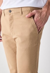 Homme portant un pantalon beige avec la main droite dans la poche avant, bouton visible et passants de ceinture, chemise blanche partiellement visible.